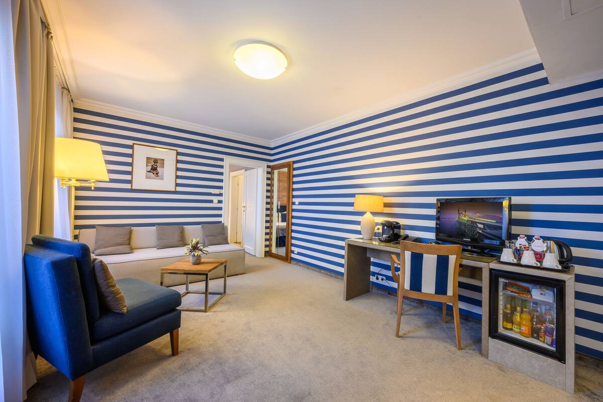 A living room with striped walls, a sofa, a chair, a coffee table, a TV, and a mini-fridge.
