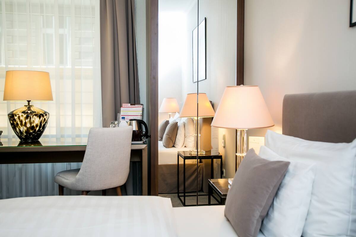 A hotel room with a bed, two bedside lamps, a desk with a lamp, a chair, and a window with curtains.