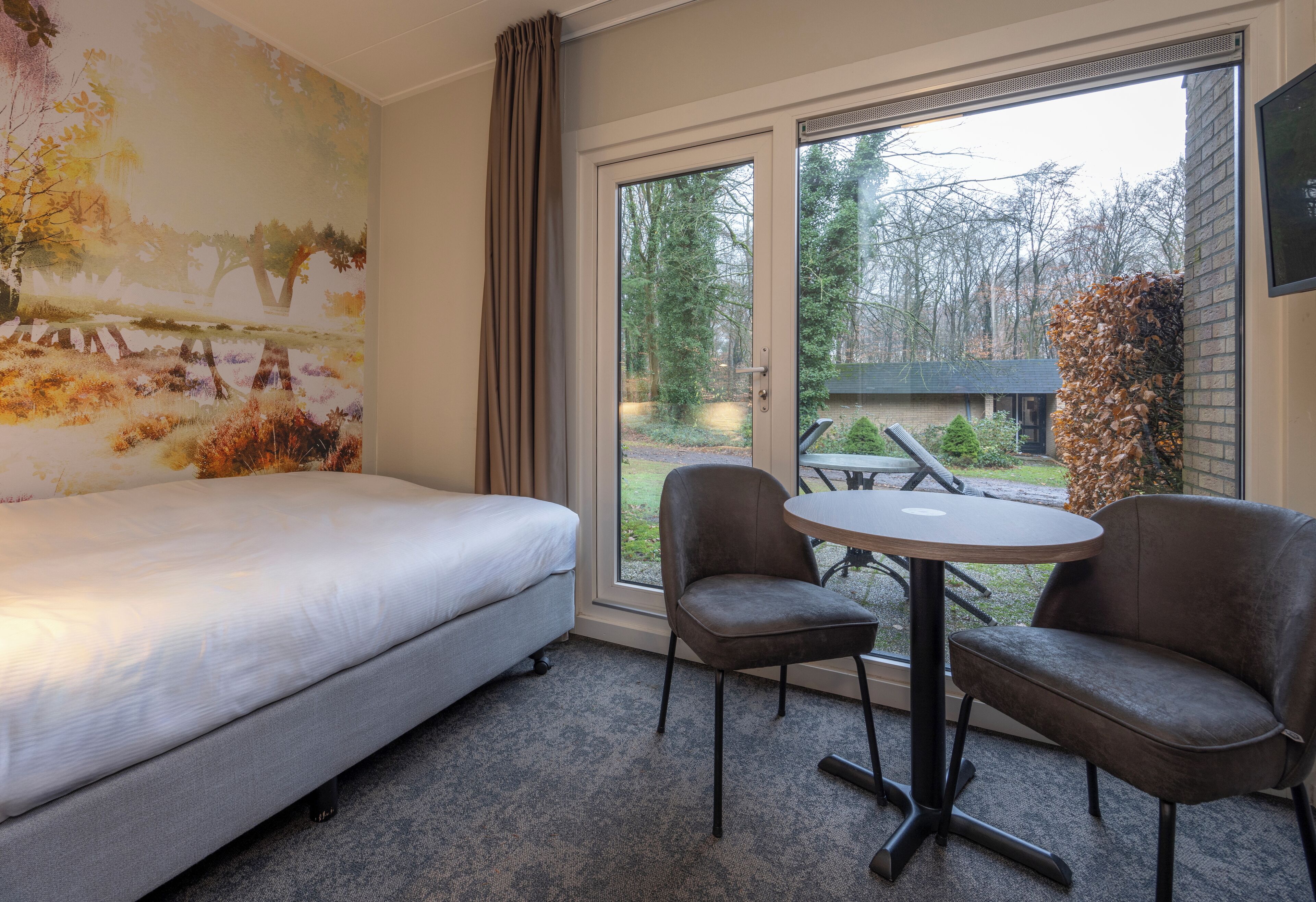 Une chambre d’hôtel avec un lit, deux chaises, une table et une grande fenêtre donnant sur des arbres et un bâtiment à l’extérieur.