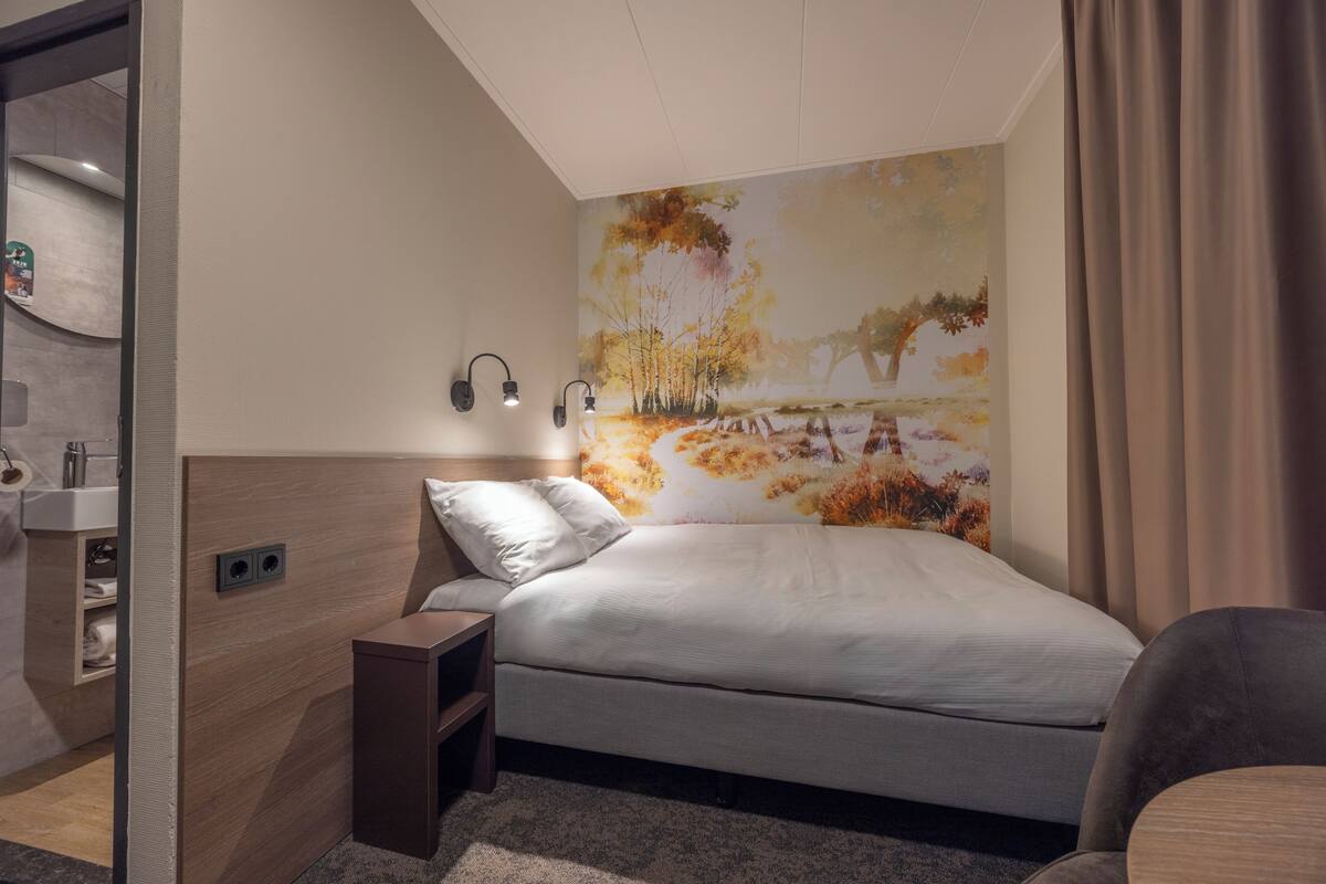 A modern hotel room with a bed, bedside table, and a wall-mounted lamp.