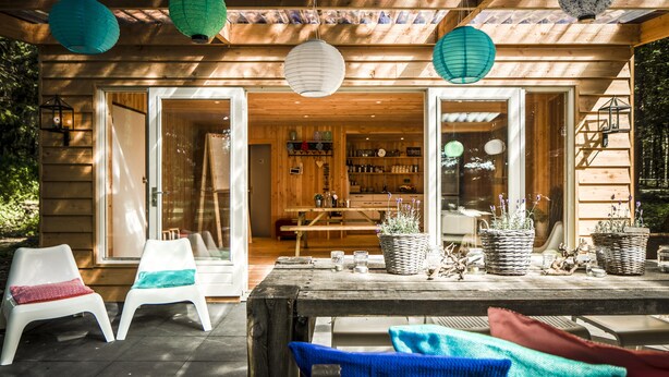 A wooden cabin with a patio area featuring two white plastic chairs, a wooden table, and potted plants. - Fletcher Hotel-Restaurant Mooi Veluwe (Putten)