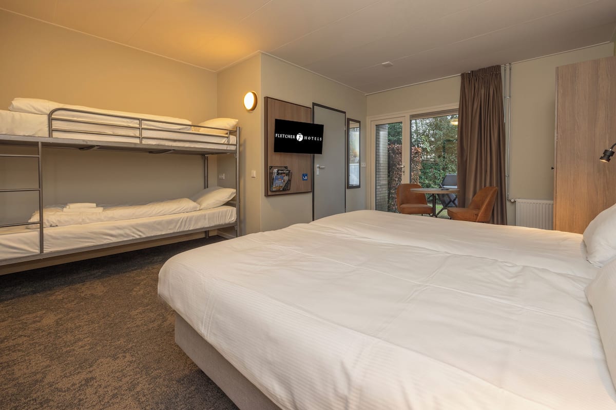 A hotel room with a large bed, a smaller bunk bed, a desk with a chair, a television, and a window with curtains.
