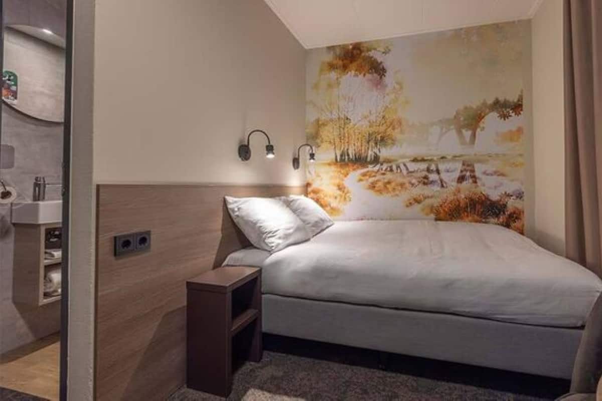 A modern bedroom with a bed, a nightstand, a wall-mounted lamp, and a large nature-themed mural.