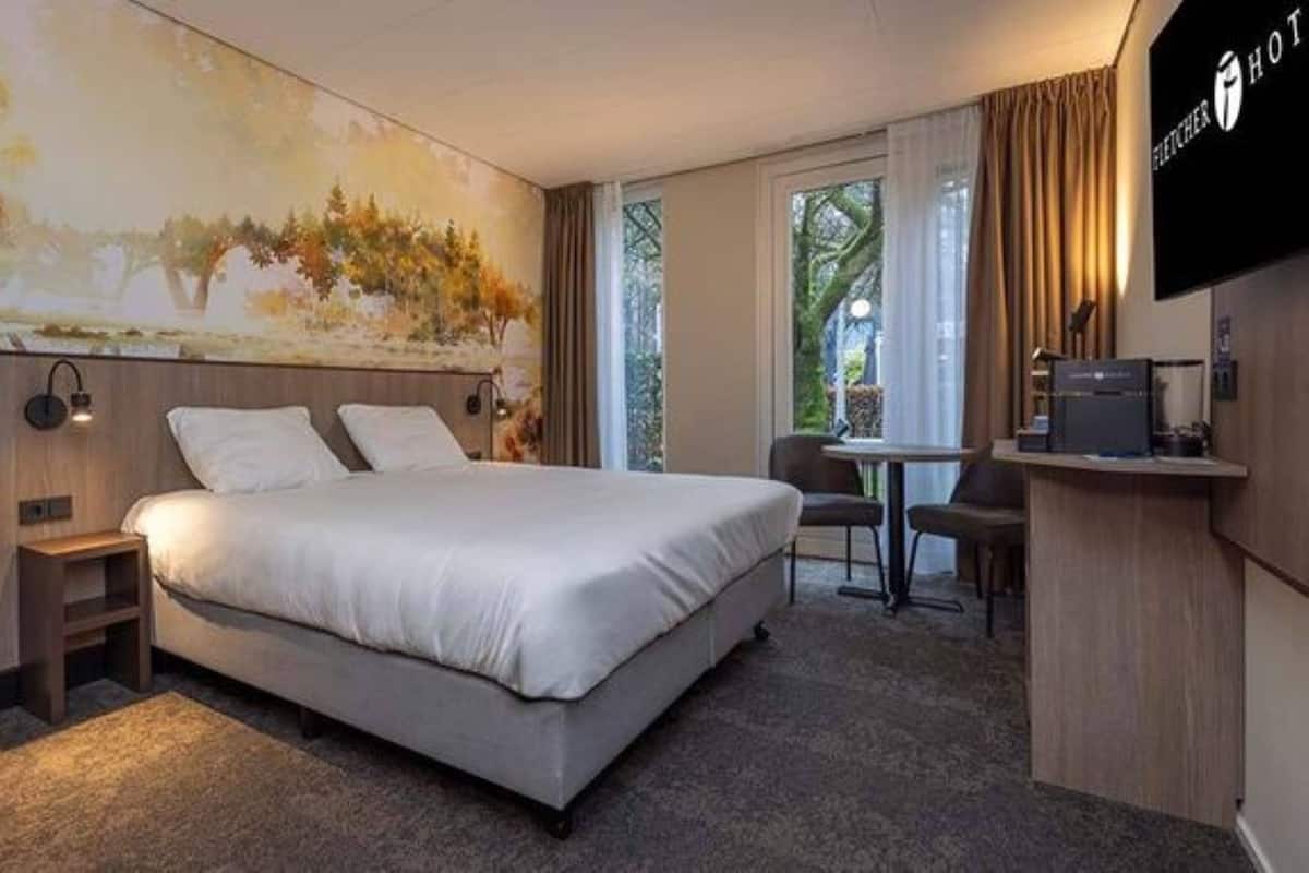 A hotel room with a bed, a desk, a chair, a TV, and a painting on the wall.