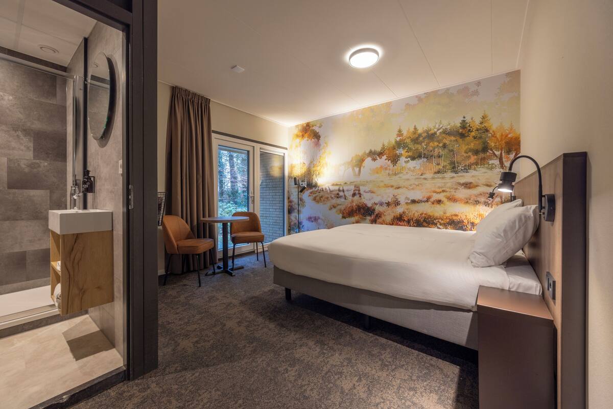 A modern hotel room with a large bed, a shower area, a small table with two chairs, and a mural on the wall.