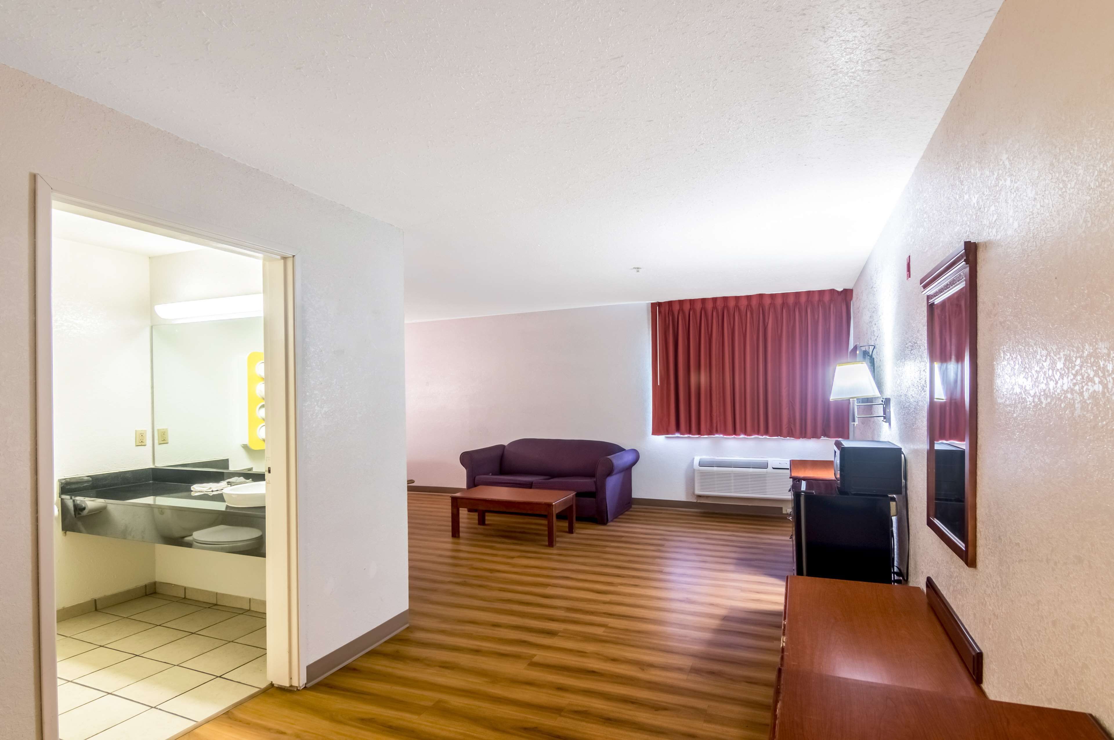 A hotel room with a wooden floor, a sofa, a TV, and a bathroom visible through an open door.