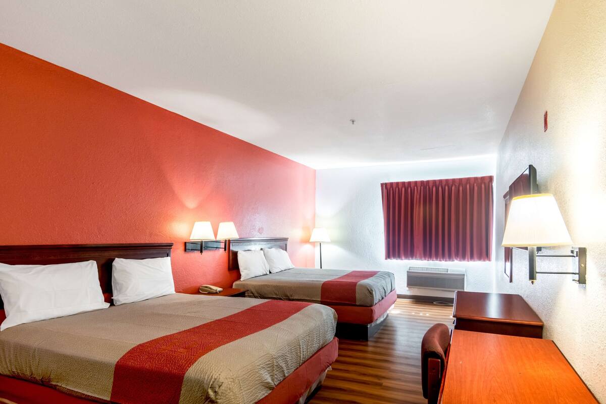 Ett hotellrum med två sängar, ett skrivbord och en stol. Det finns två väggmonterade lampor och ett fönster med gardiner.
