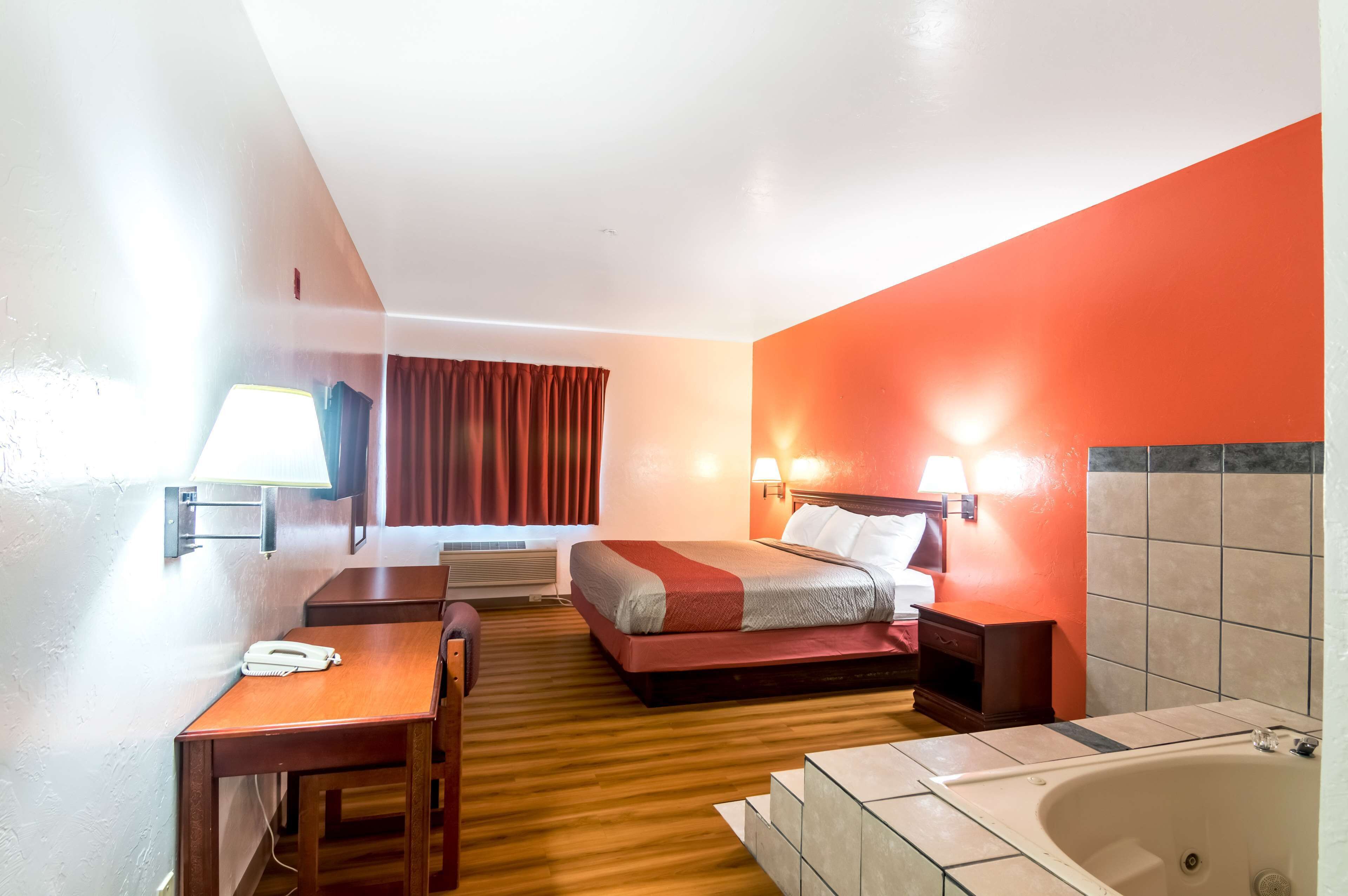 A hotel room with a bed, a desk, a telephone, a lamp, a window with curtains, a wall-mounted lamp, a bathtub, and a tiled wall.