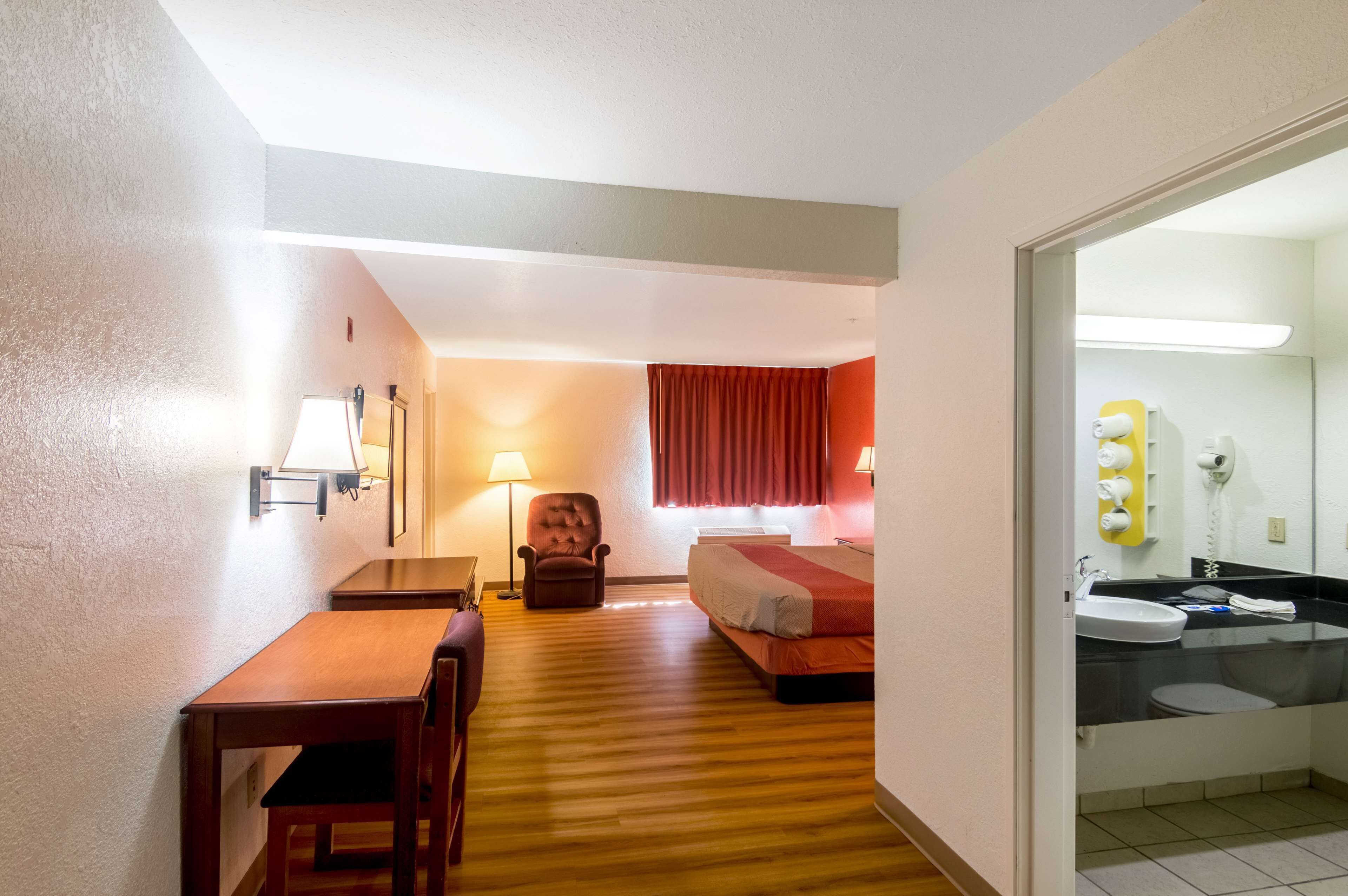A hotel room with a wooden floor, a bed, a chair, a desk, and a bathroom with a sink and mirror.