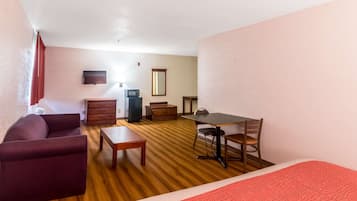 A compact room with a sofa, a small table, a desk with a chair, a television, and a mirror.