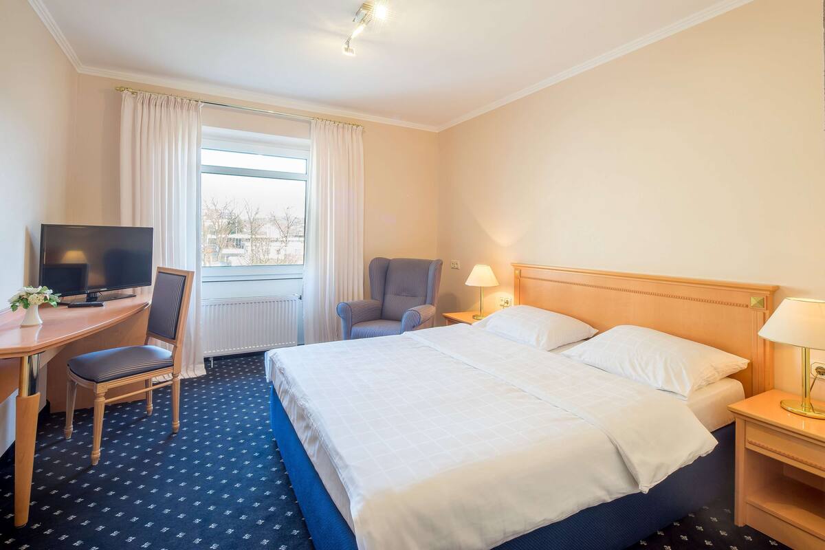 Une chambre d’hôtel avec un lit, un bureau avec une télévision, une chaise, une lampe et une fenêtre avec des rideaux.