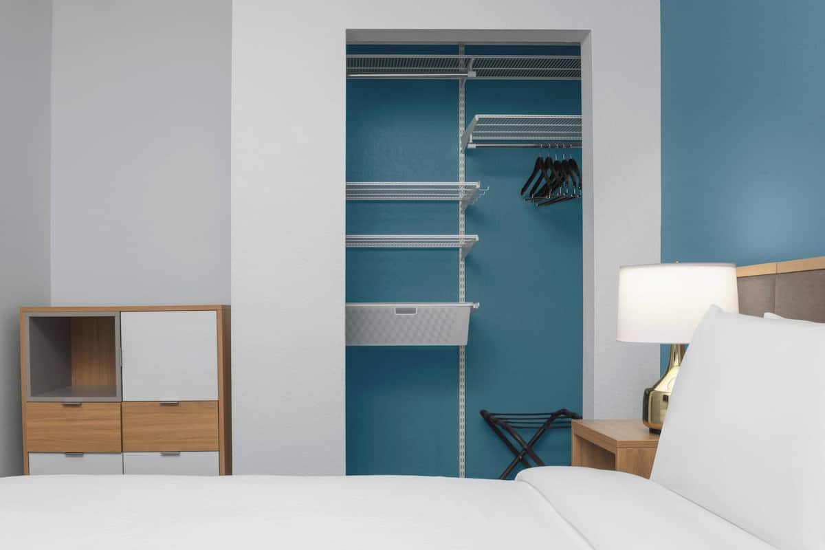 Una habitación de hotel con cama, mesita de noche con lámpara, cómoda de madera y armario con estantes y ropa colgada.