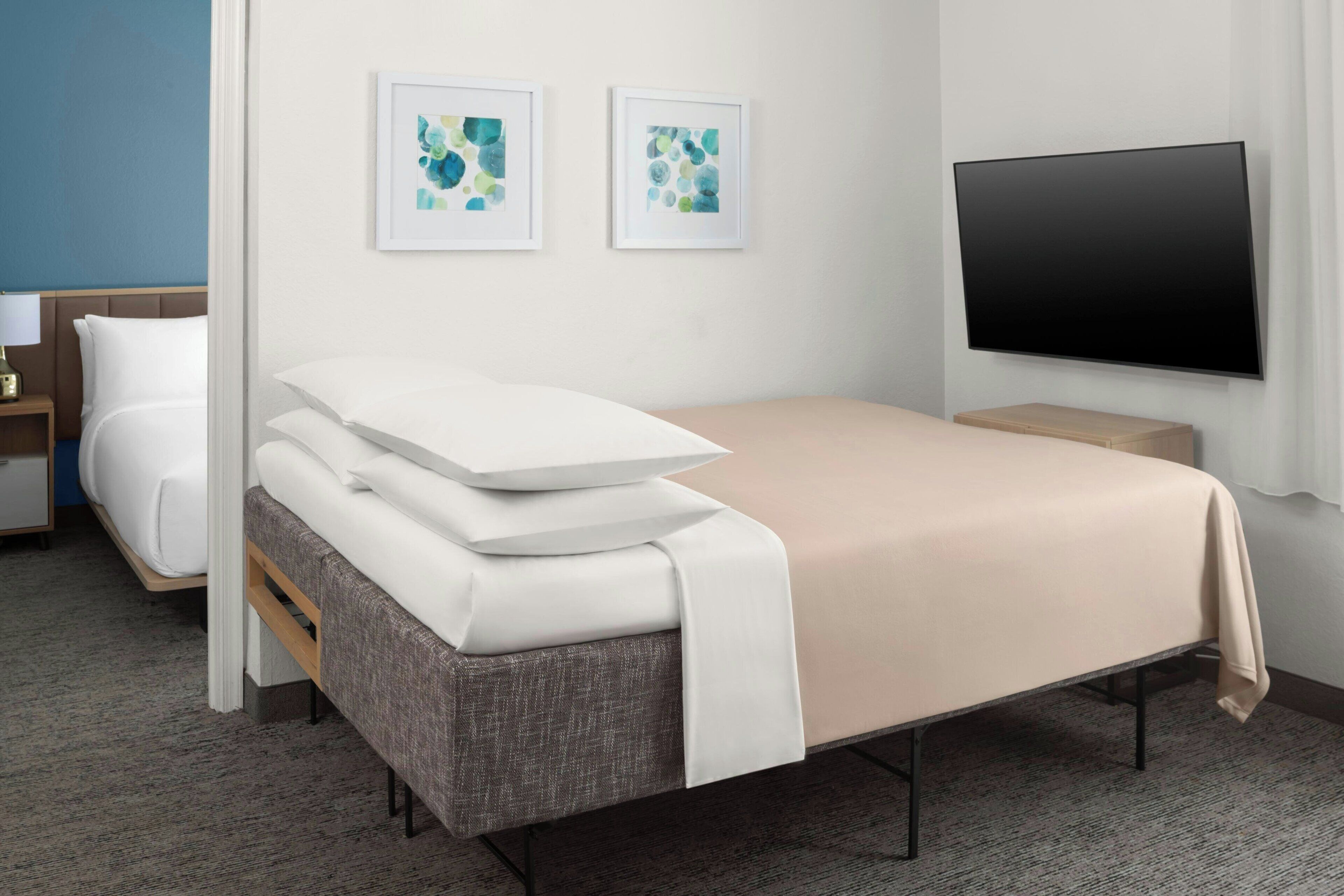 A hotel room with a bed, a TV, two framed artworks, and a bedside table.