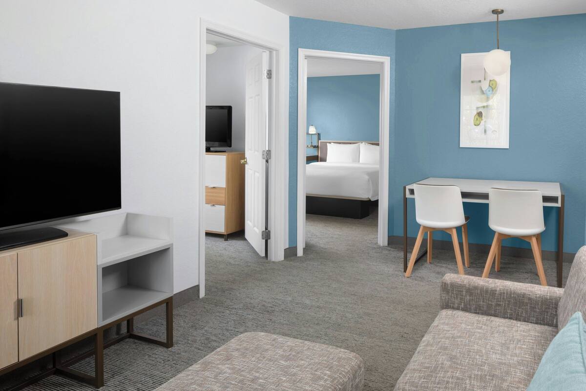 Habitación de hotel con televisión, un escritorio con dos sillas, una cama y una puerta que da a otra habitación.