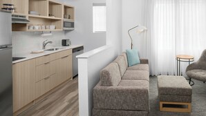 A modern kitchen with wooden cabinets, a built-in microwave, and a countertop. There is a sofa and ottoman in the living area, a small round table, and a lamp.