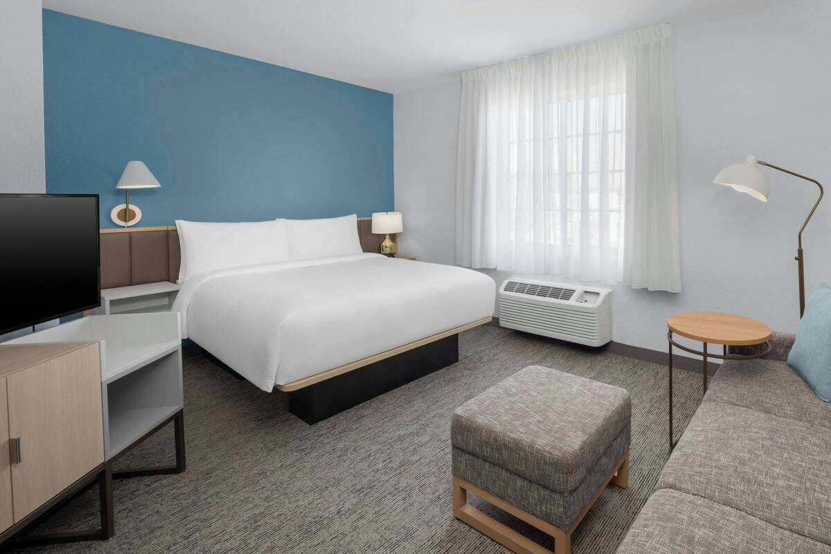 Habitación de hotel con cama, televisor, escritorio, silla, sofá, lámpara y ventana con cortinas.