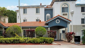 A multi-story building with a tiled roof, a central entrance with a 'WELCOME' sign, and a well-maintained garden with various shrubs and a palm tree.