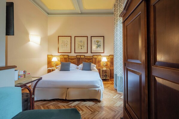 A hotel room with a large bed, two bedside tables with lamps, a wooden wardrobe, and three framed botanical prints on the wall.
