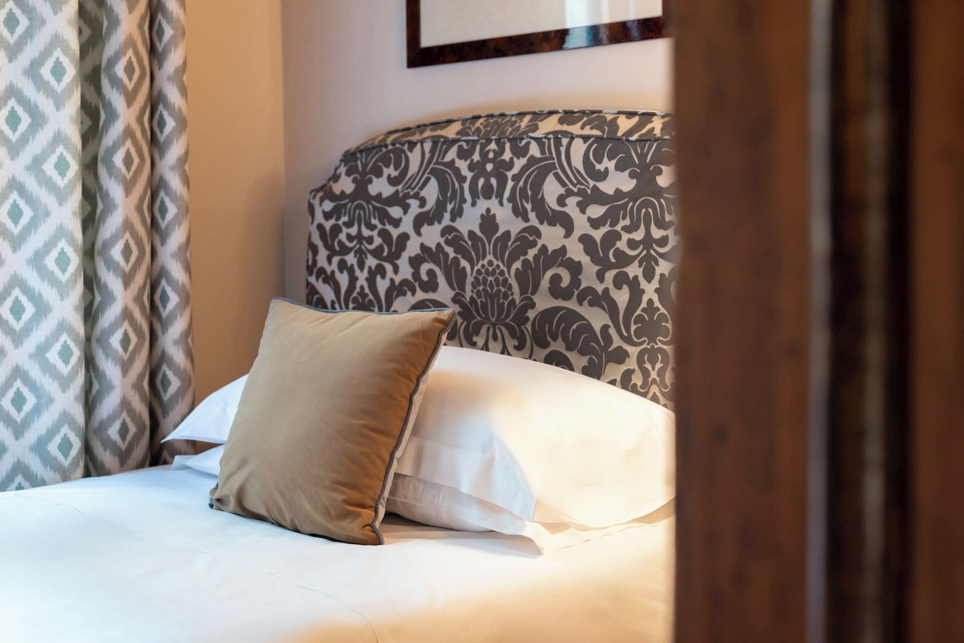 Un letto rifatto con cura, una testiera a fantasia, un cuscino beige e uno bianco.