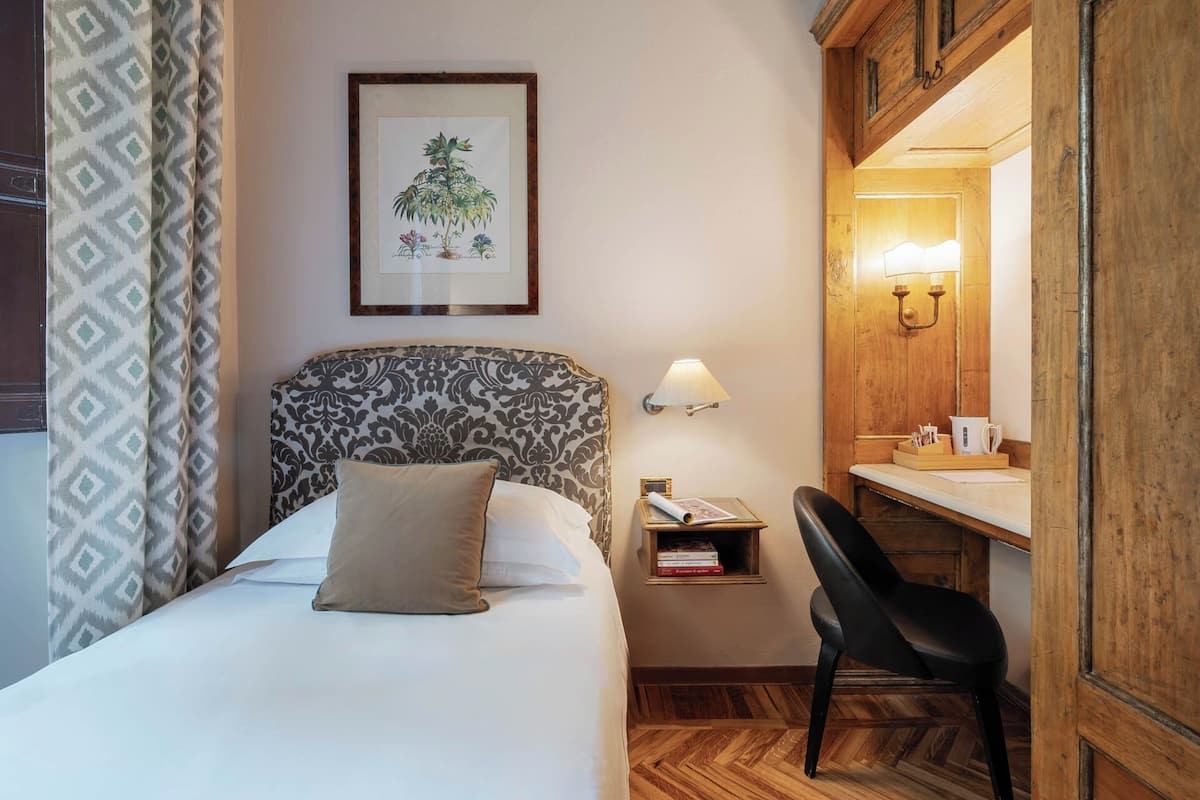 A hotel room with a bed, a desk, a chair, a nightstand with a lamp, and a framed botanical illustration on the wall.