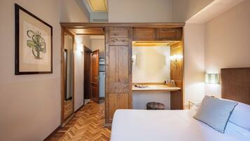 Habitación de hotel con piso de madera, cama con ropa de cama blanca, armario de madera, espejo e impresión enmarcadas de motivos botánicos en la pared.