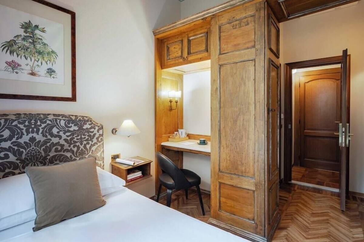 A hotel room with a bed, a desk, a chair, a wardrobe, and a framed botanical illustration on the wall.