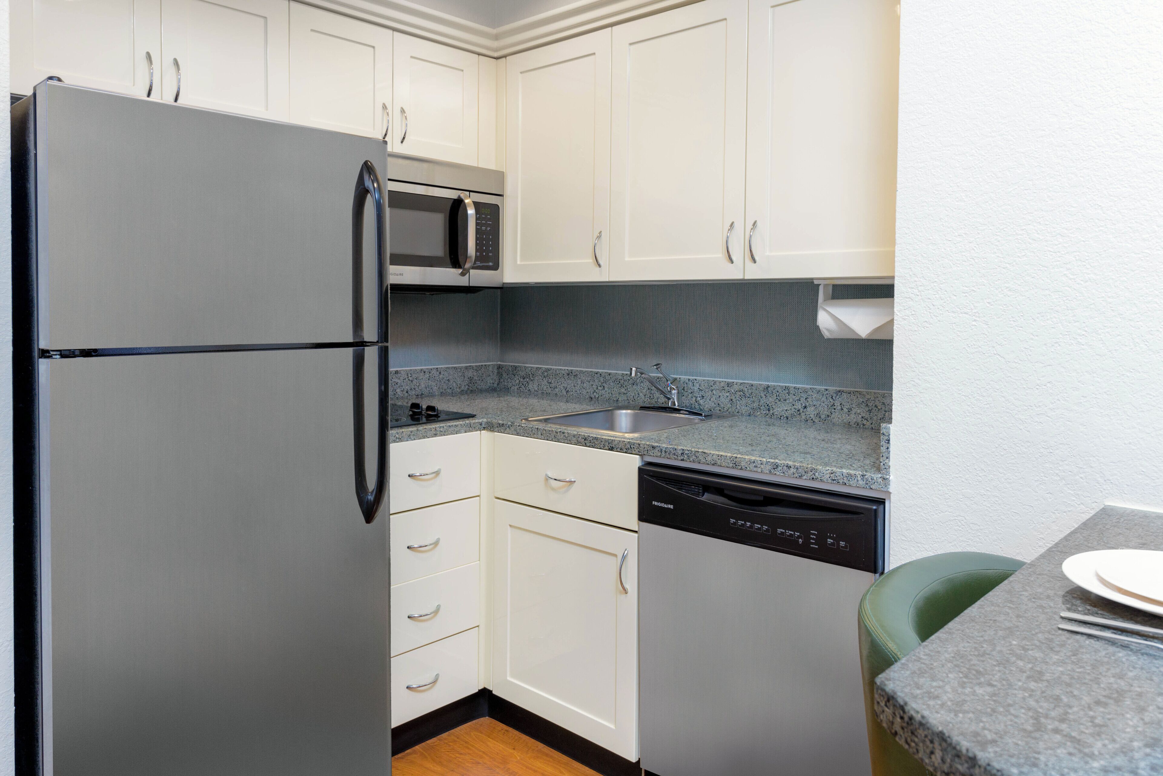 A compact kitchen with white cabinets, a stainless steel refrigerator, a microwave, a dishwasher, and a granite countertop.