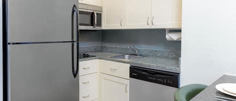 A compact kitchen with white cabinets, a stainless steel refrigerator, a microwave, a dishwasher, and a granite countertop.