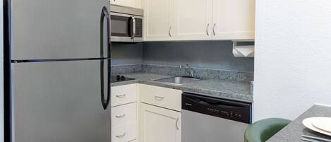 A compact kitchen with white cabinets, a stainless steel refrigerator, a microwave, a dishwasher, and a granite countertop.