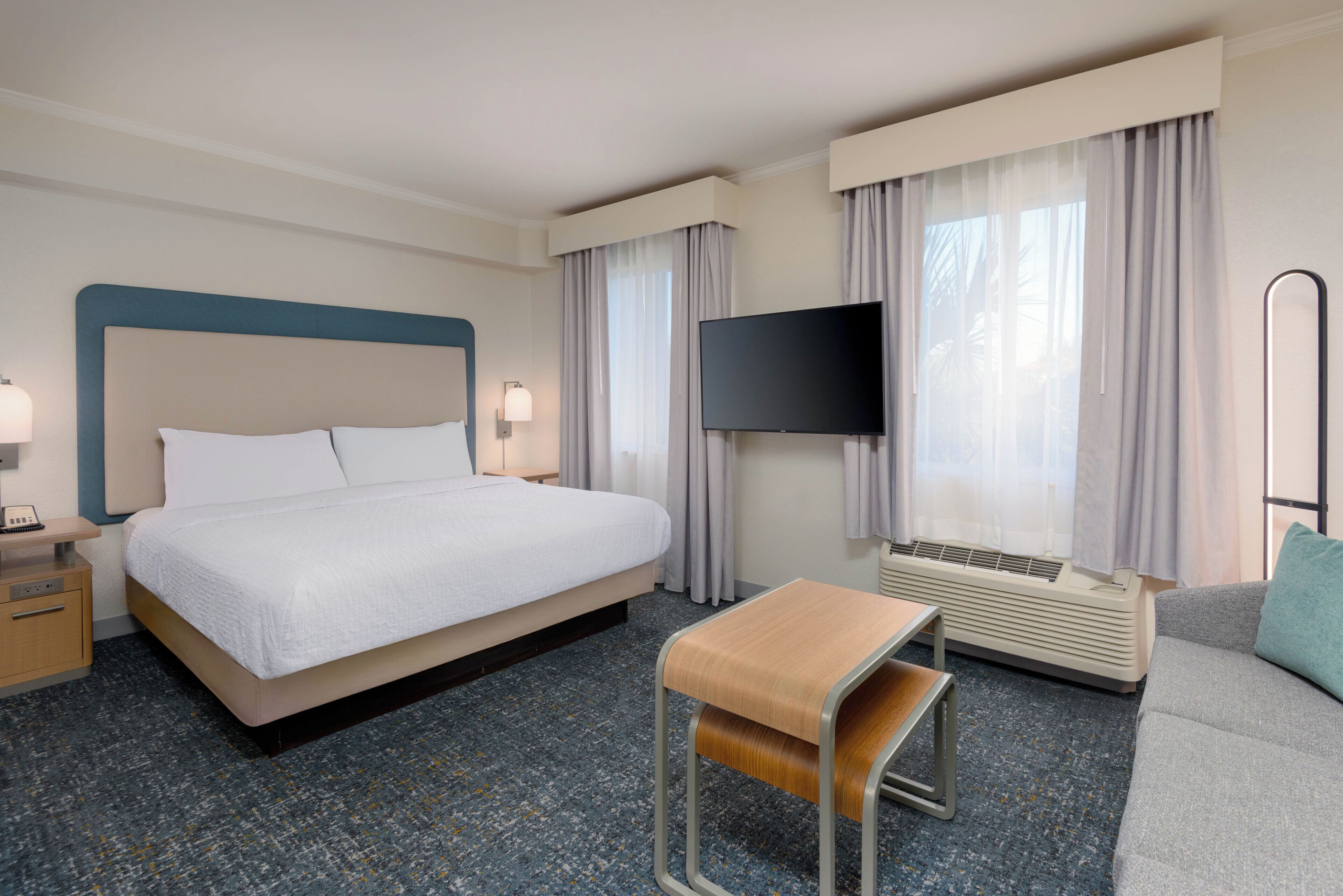 Quarto de hotel com cama, mesa de cabeceira, luminária, televisão, aparelho de ar condicionado e uma pequena mesa com um banco.