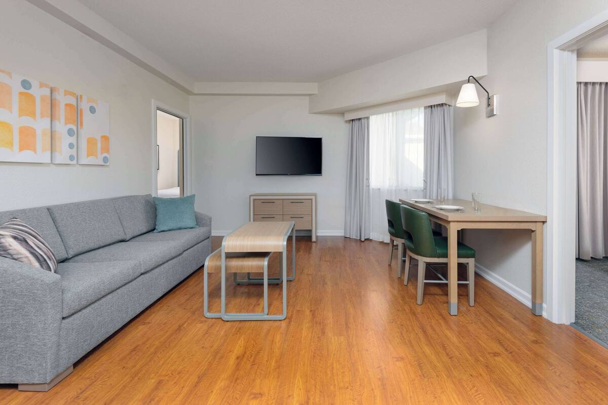 Ein modernes, gut beleuchtetes Zimmer mit einem grauen Sofa, einem Holzespeisetisch mit zwei Stühlen, einem an der Wand montierten Flachbildfernseher und einem kleinen Holznachttisch.