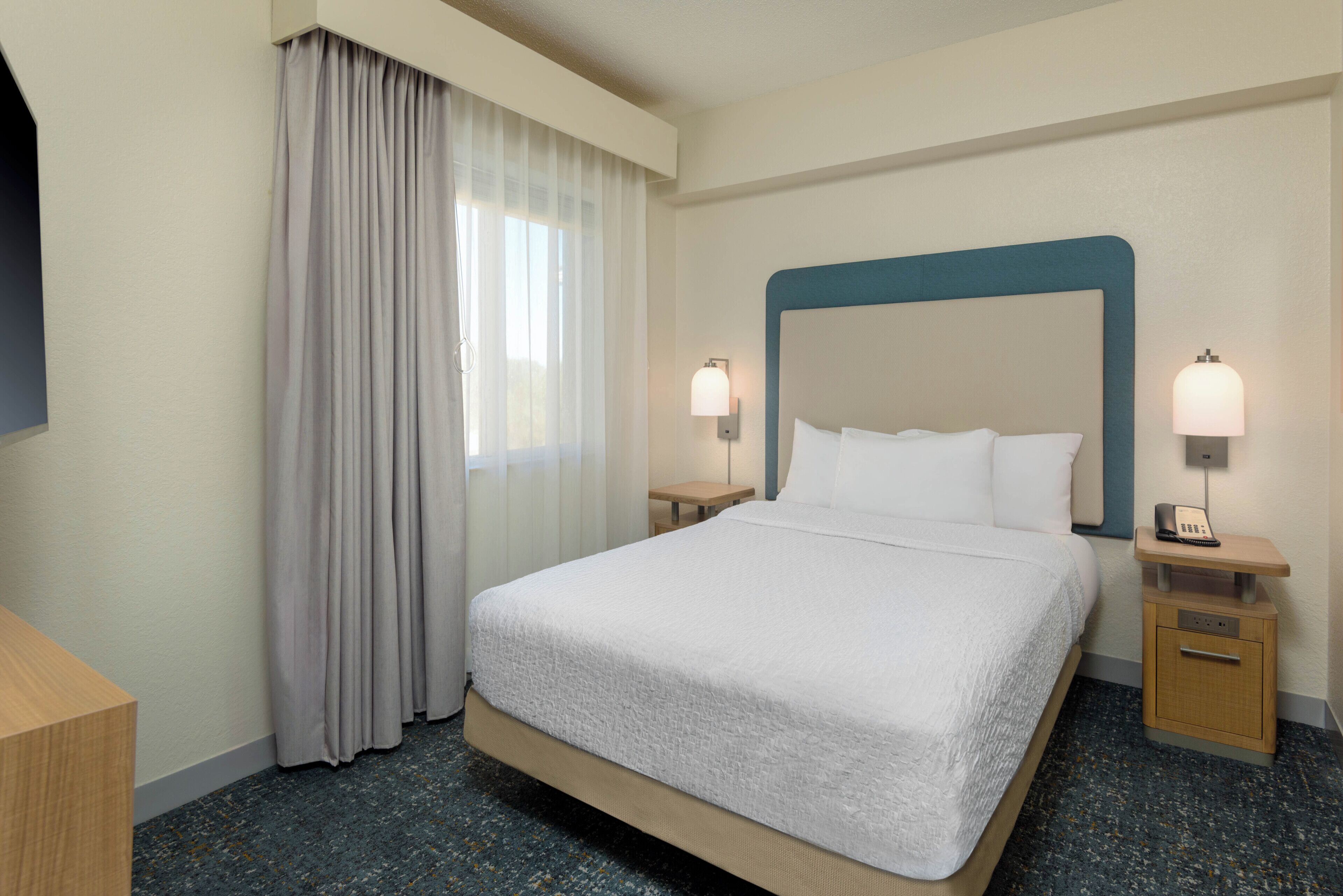 A hotel room with a bed, two bedside tables, a nightstand, a wall-mounted telephone, and a window with curtains.