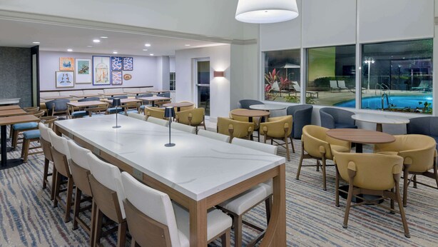 A modern hotel lobby with a long communal table, several smaller tables, and chairs. There are framed artworks on the walls, a pool area visible through large windows, and a carpeted floor. - Homewood Suites by Hilton Bonita Springs Naples North (Bonita Springs)