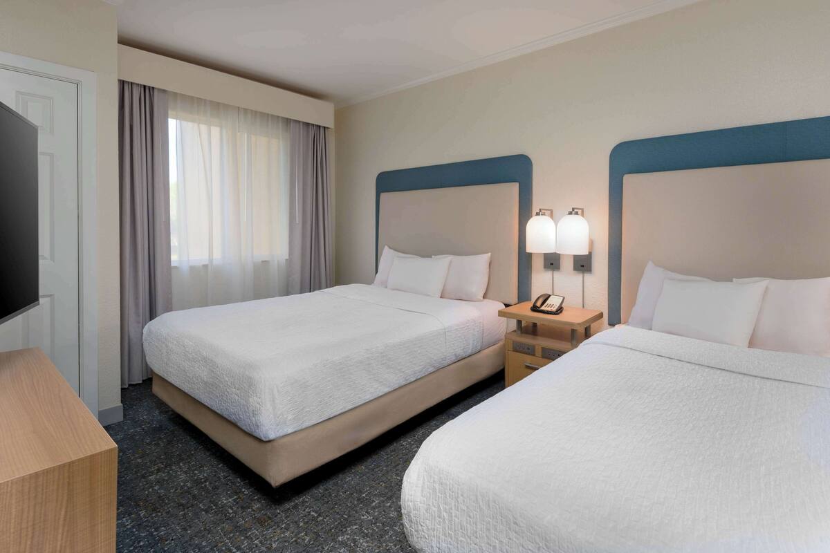 Ein Hotelzimmer mit zwei Betten, einem Fernseher, einem Schreibtisch und einem Fenster mit Vorhängen.