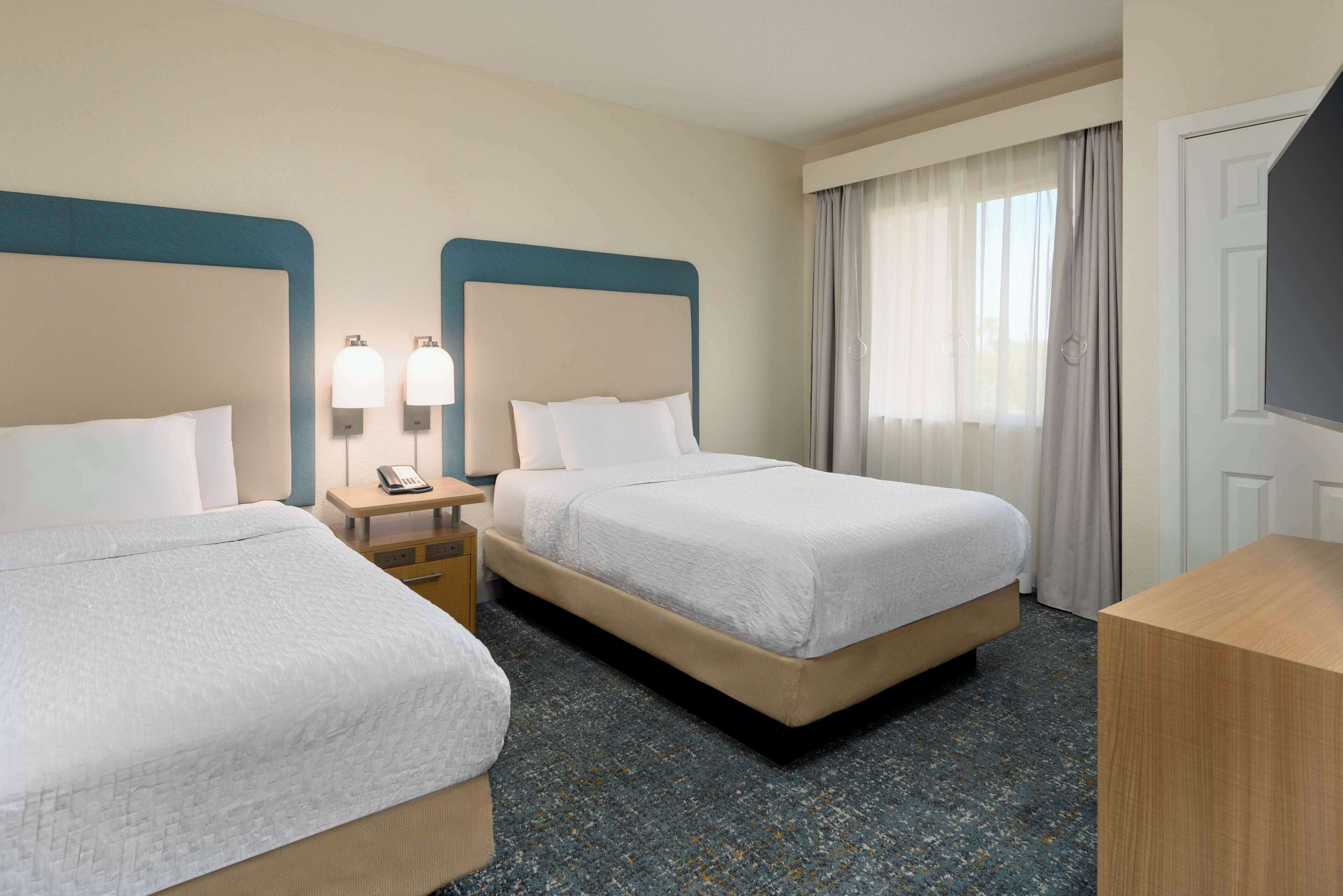 A hotel room with two beds, a nightstand with a phone, and a large window with curtains.