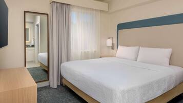 Una habitación de hotel con una cama grande, una mesita de noche de madera, un espejo y una ventana con cortinas traslúcidas.