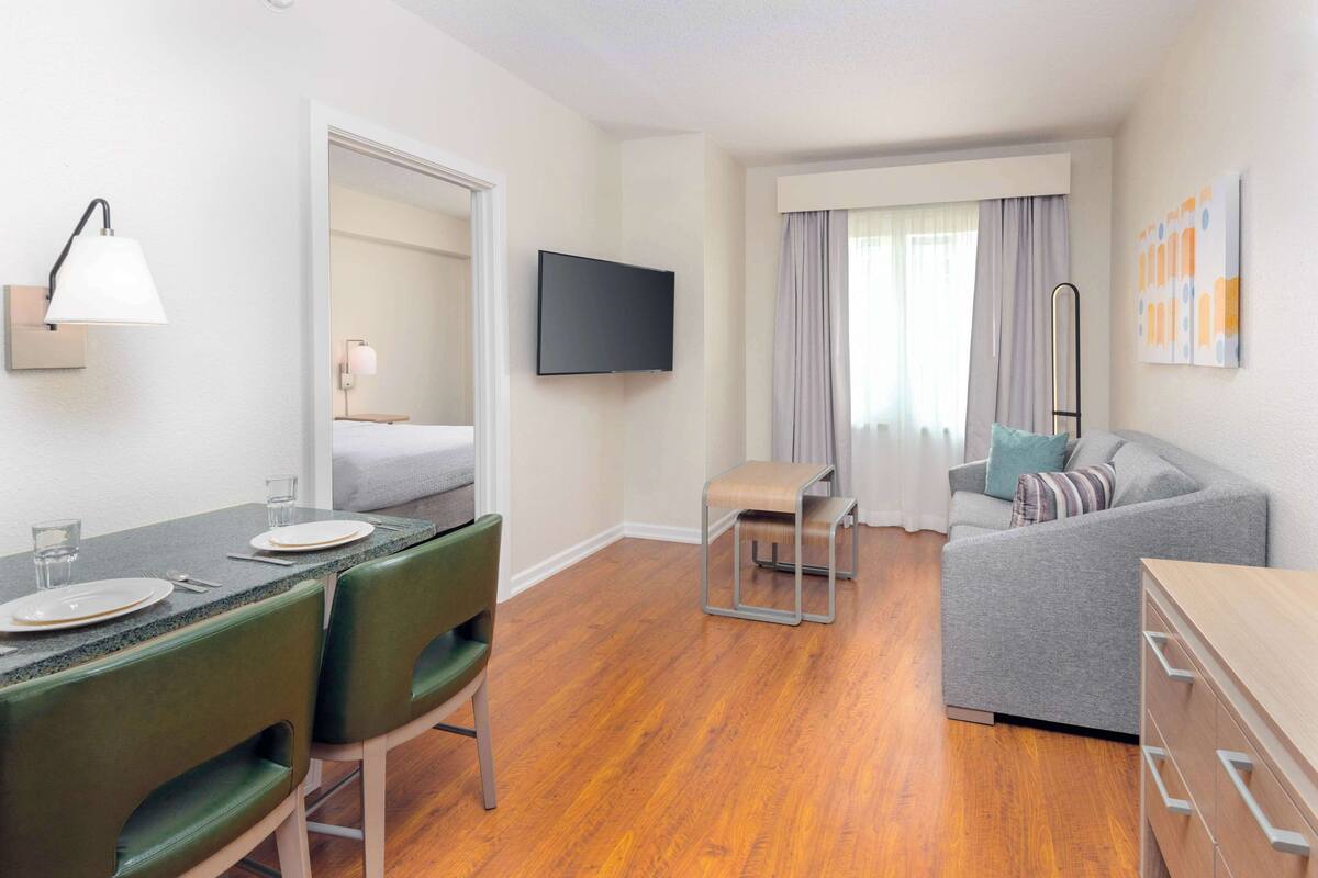 Ein modernes Hotelzimmer mit Essbereich, einer Couch, einem Fernseher und einem Fenster mit Vorhängen.
