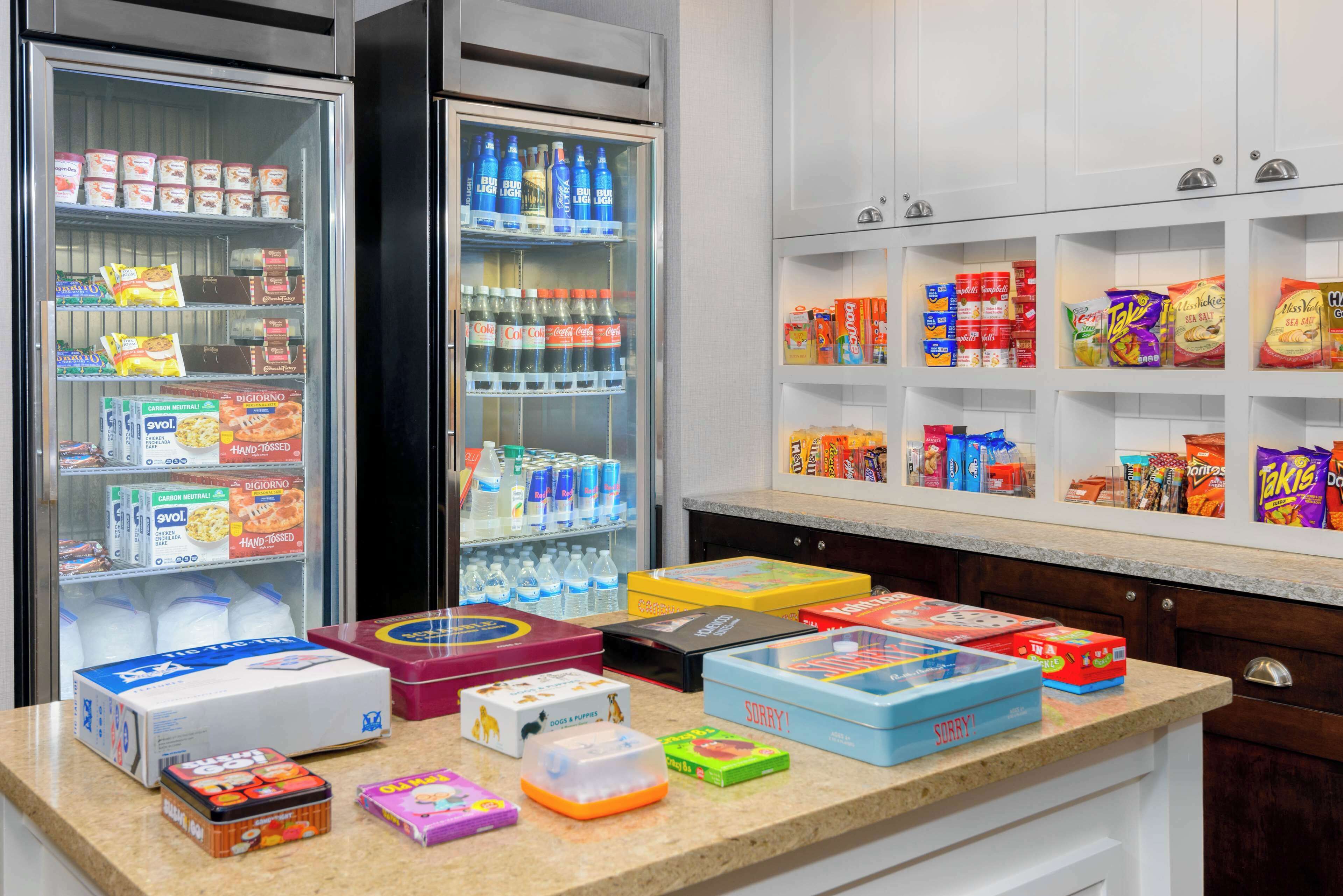 A kitchen with a refrigerator stocked with beverages and snacks, a countertop with various food items, and cabinets above the counter.