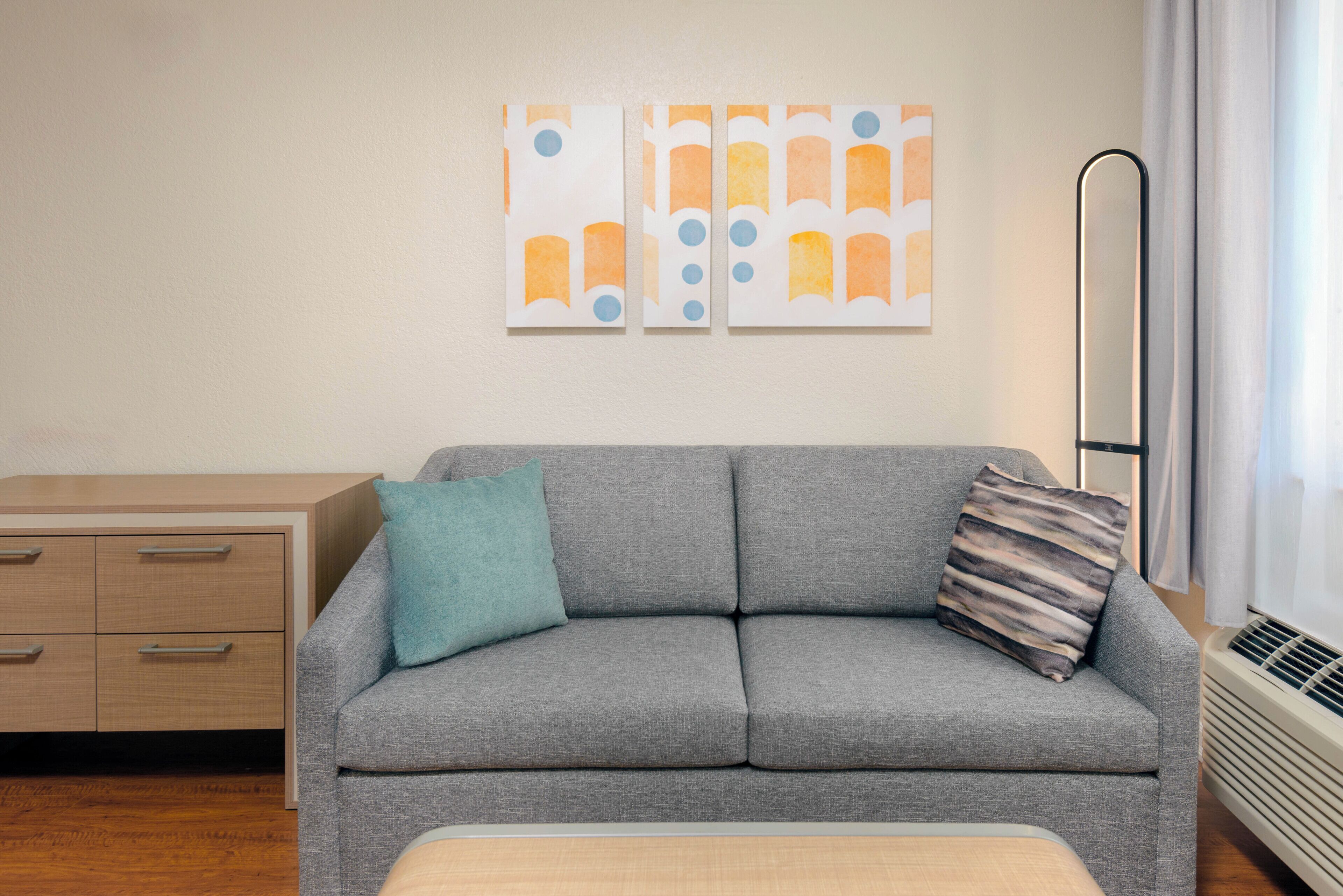 Un canapé gris avec deux coussins, une table d’appoint en bois et un mur orné d’œuvres d’art abstraites.