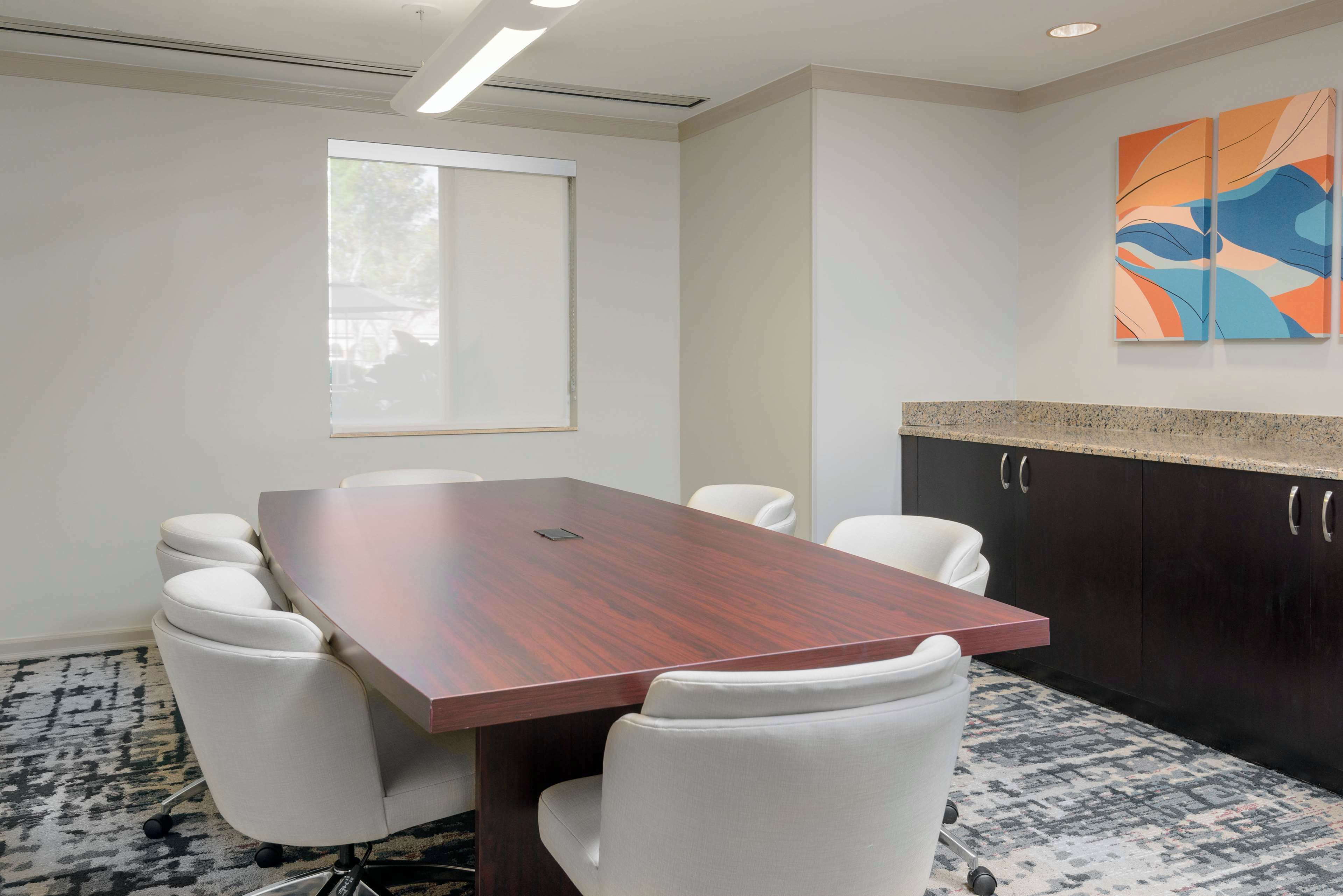A conference room with a large wooden table, white chairs, a window, and abstract wall art.
