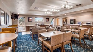 A hotel dining area with multiple tables and chairs, a kitchen area with appliances, and a television mounted on the wall. - Best Western Plus Gateway Inn & Suites (Aurora)