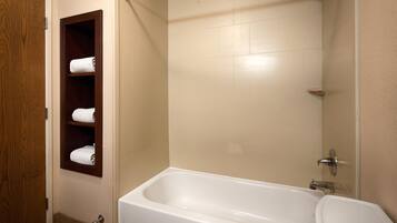Une salle de bain avec une baignoire blanche, des murs recouverts de carreaux beiges et une étagère en bois où sont posées des serviettes.