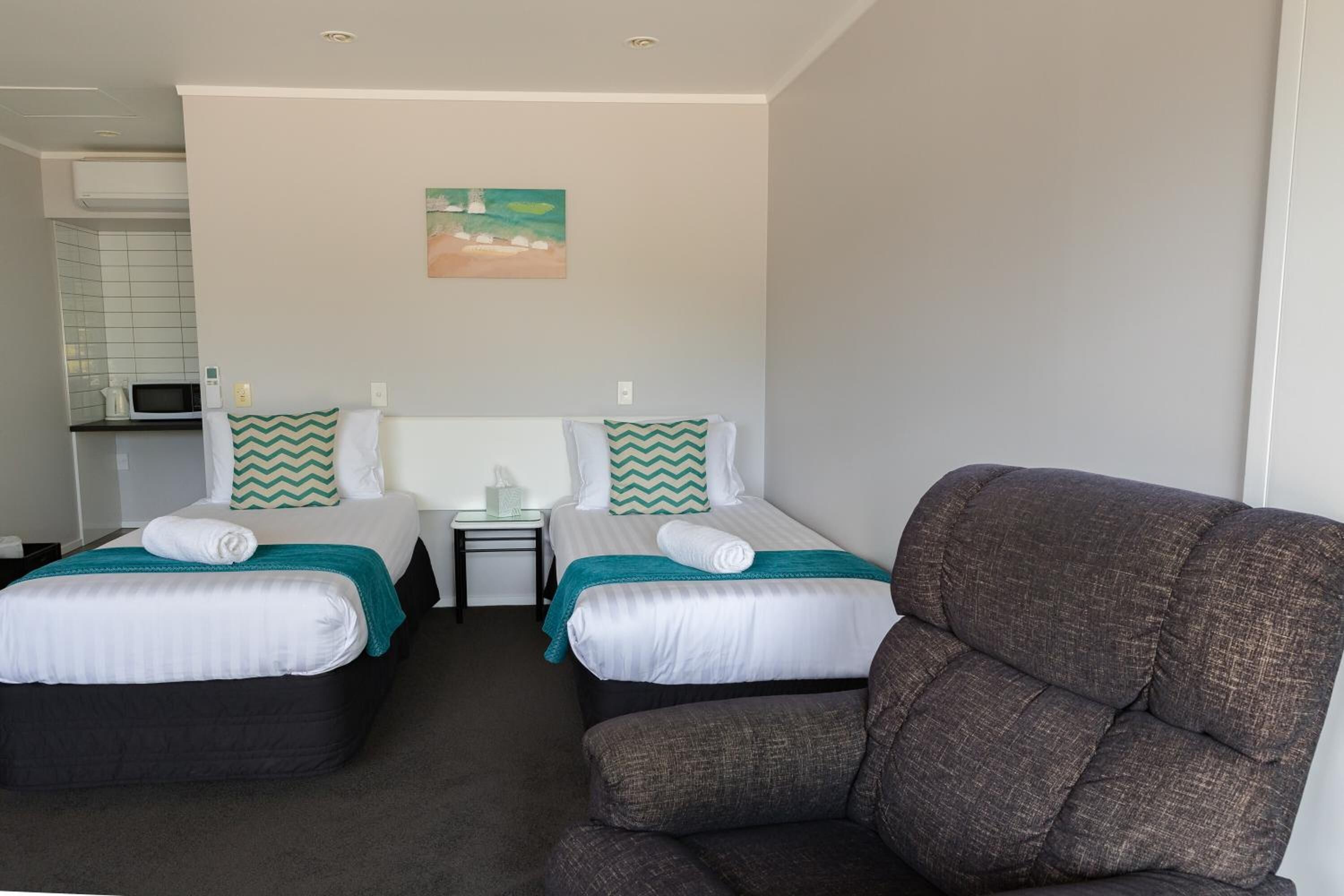 A hotel room with two beds, a gray armchair, a small table with a lamp, and a painting on the wall.