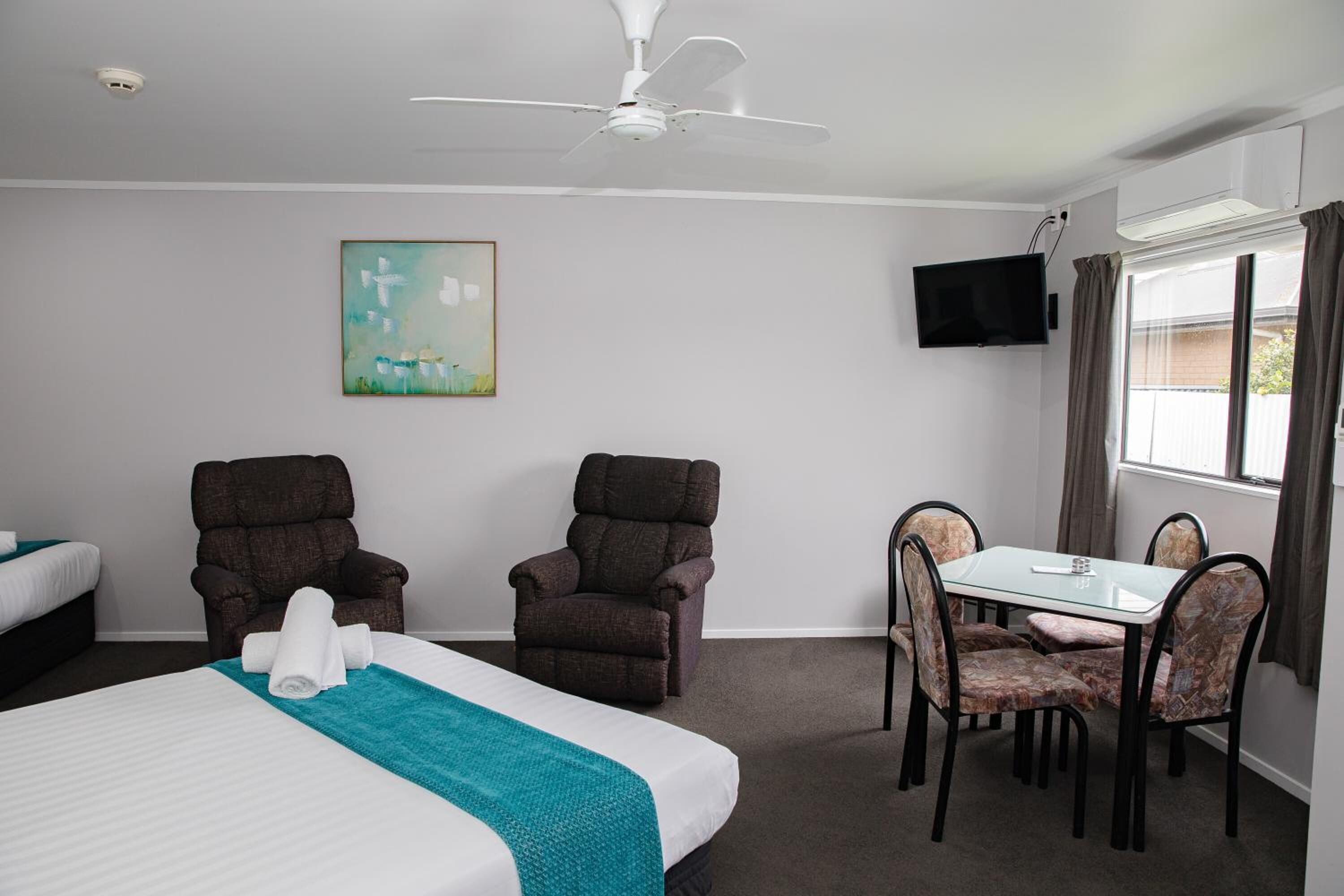 A hotel room with a bed, two armchairs, a dining table with chairs, a ceiling fan, a painting, a TV, and a window with curtains.