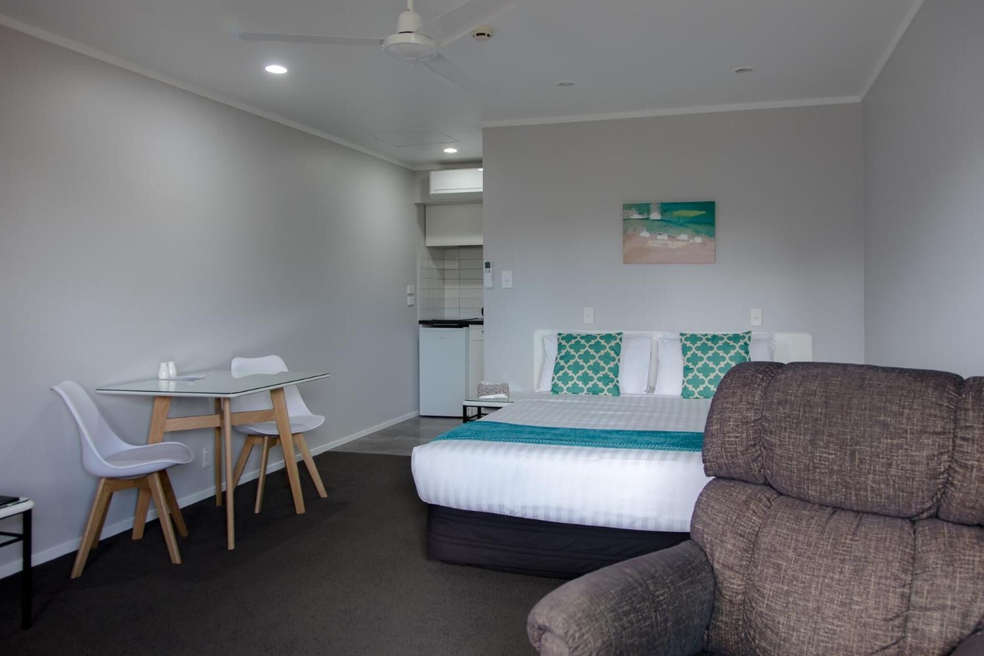 Ein kleines Hotelzimmer mit einem Bett, einem Schreibtisch und einem Stuhl. Im Hintergrund ist eine Küchenzeile zu sehen.