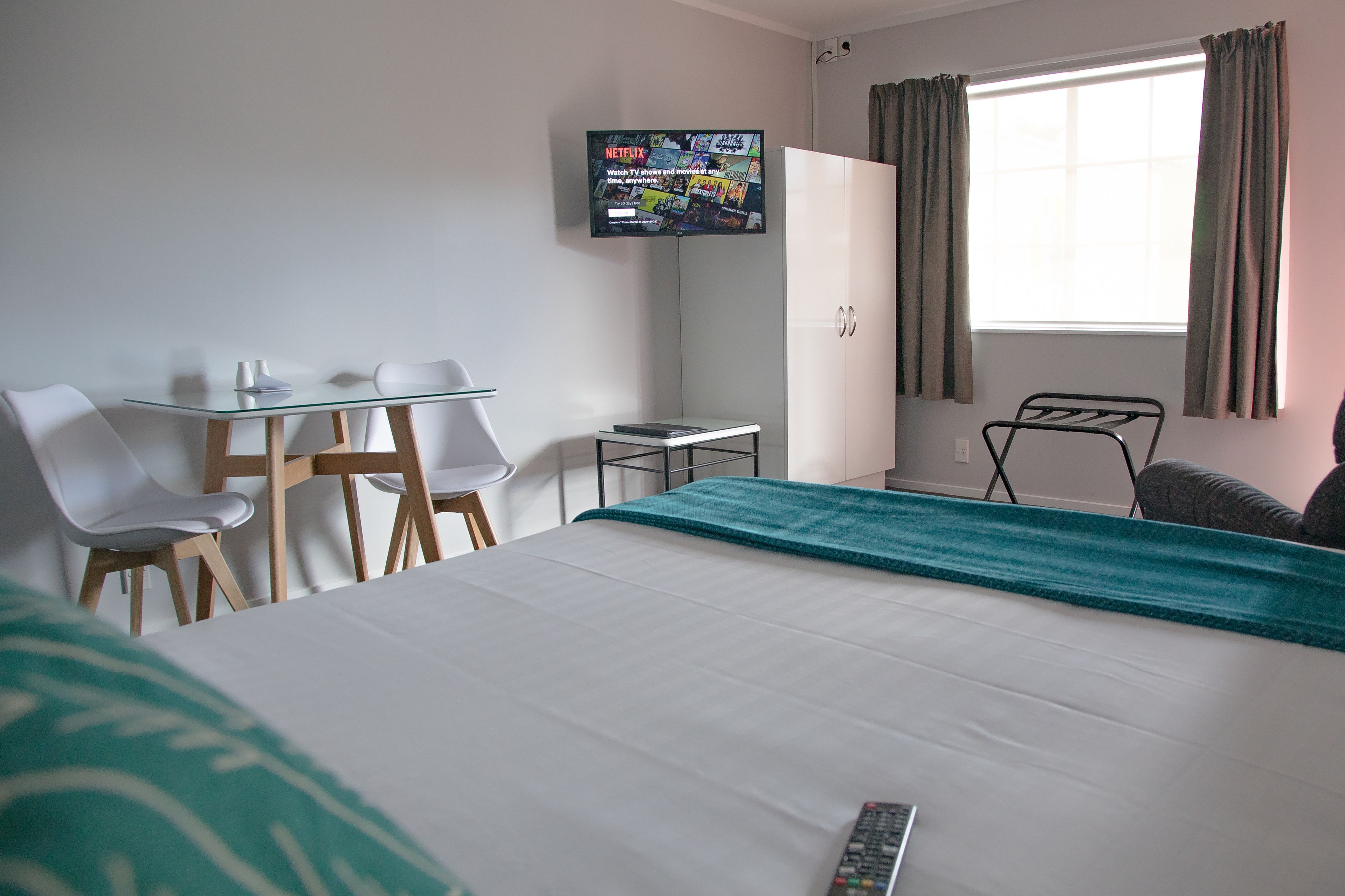 A hotel room with a bed, a desk with chairs, a TV mounted on the wall, and a window with curtains.