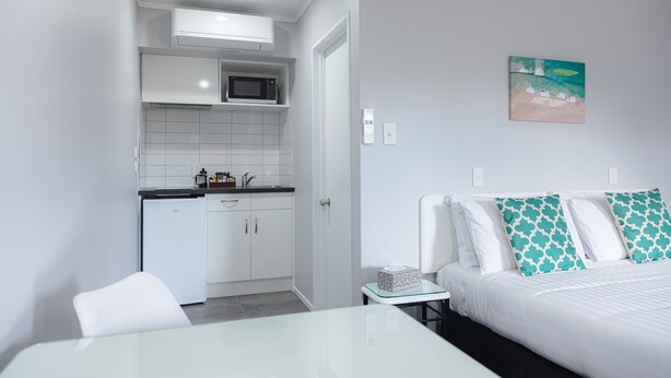 A compact hotel room with a kitchenette, a bed with patterned pillows, and a small bedside table. - Bk's Palm Court Motor Lodge (Gisborne)