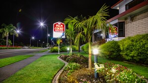 A well-lit hotel entrance with a palm tree and flower beds, a clear road, and a 'Palm Court' sign. - Bk's Palm Court Motor Lodge (Gisborne)