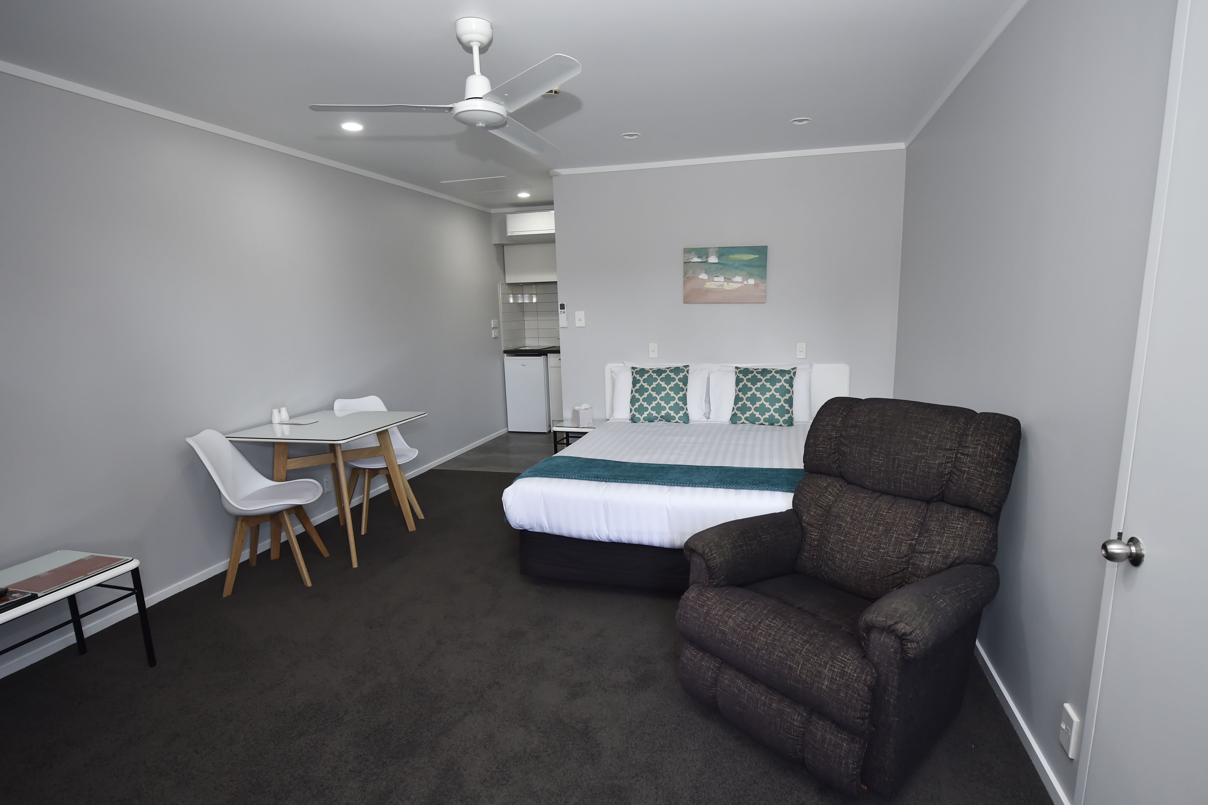 A bedroom with a bed, a desk, chairs, a ceiling fan, and a painting on the wall.