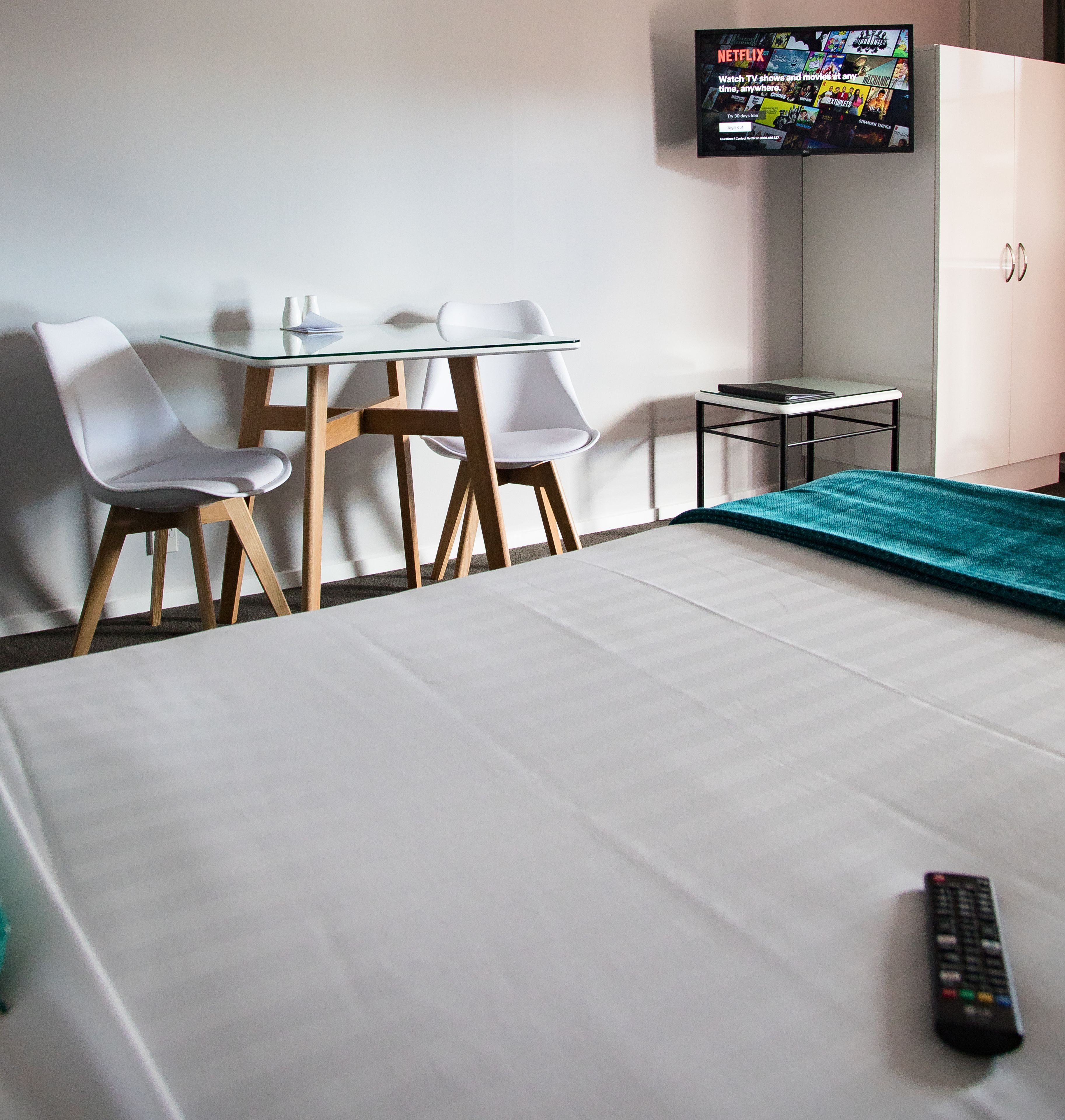 Une chambre d’hôtel avec un lit, un téléviseur fixé au mur, une petite table, deux chaises et une télécommande posée sur le lit.